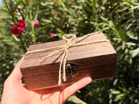 A hand holding the closed wooden keepsake box, tied with a jute twine bow, in front of green foliage.
